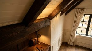 Cozy attic reading nook featuring dark wood beams, built-in wooden desk, and a brass lamp under a sloped ceiling.