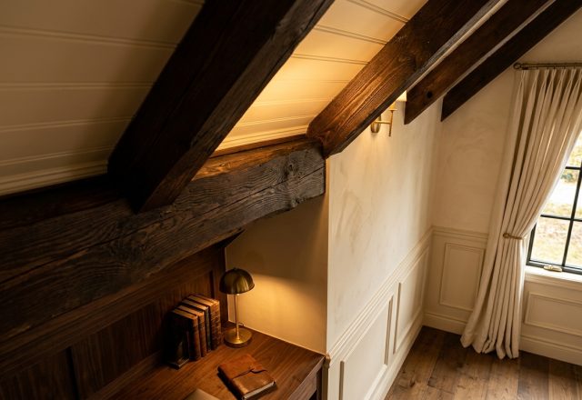 Cozy attic reading nook featuring dark wood beams, built-in wooden desk, and a brass lamp under a sloped ceiling.
