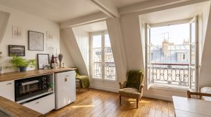 Bright attic studio apartment with sloped ceilings, wooden floors, and a compact kitchenette next to dormer windows.