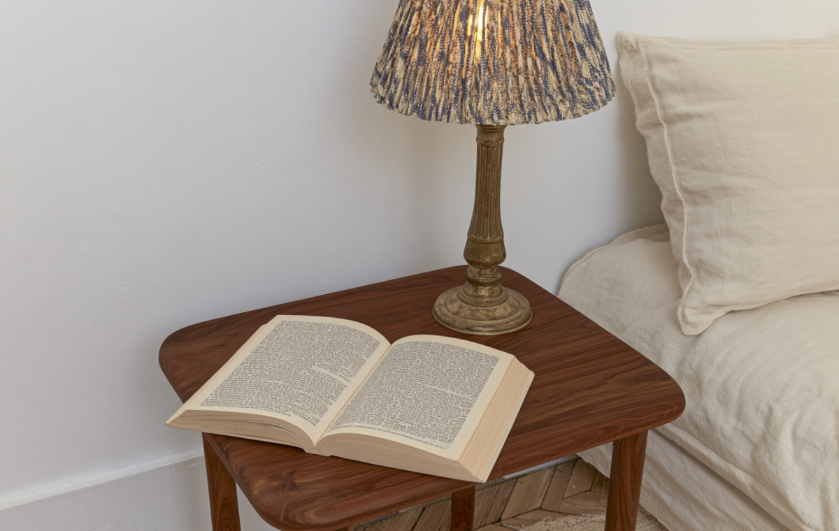 A cozy bedside reading nook featuring a walnut nightstand, open book, and a textured lamp next to a neutral bed.