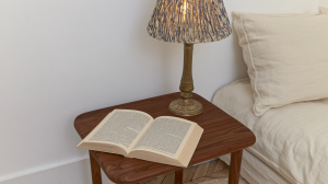 A cozy bedside reading nook featuring a walnut nightstand, open book, and a textured lamp next to a neutral bed.