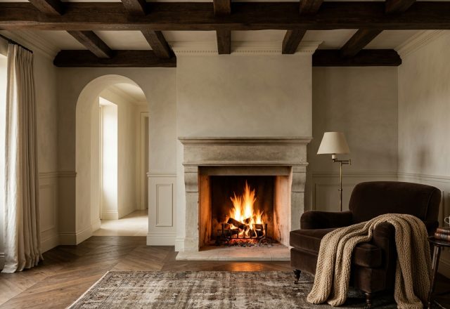 A rustic living room fireplace featuring exposed wood beams, limewash walls, and a cozy velvet armchair.