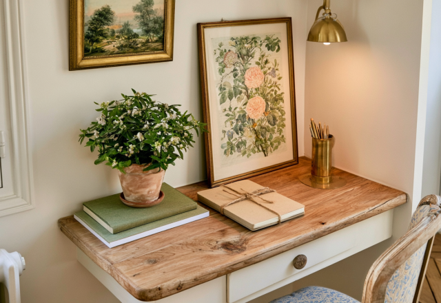 A cozy small home office nook featuring a wooden desk, vintage floral art, and a brass lamp.