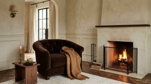 A warm neutral fireplace living room featuring a brown velvet armchair and herringbone wood floors.