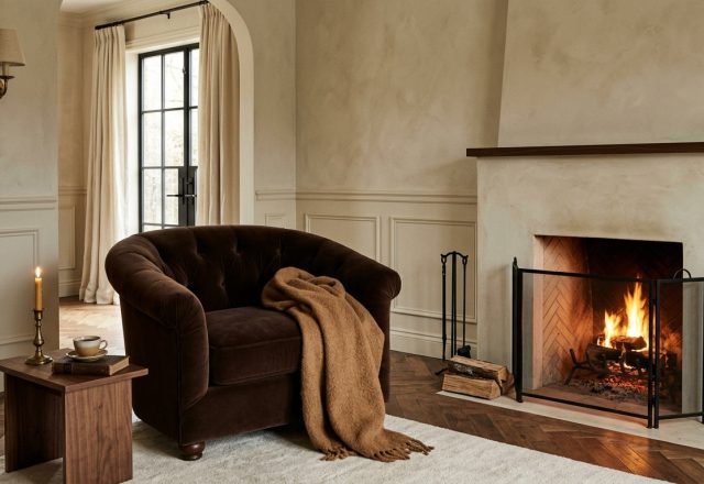 A warm neutral fireplace living room featuring a brown velvet armchair and herringbone wood floors.