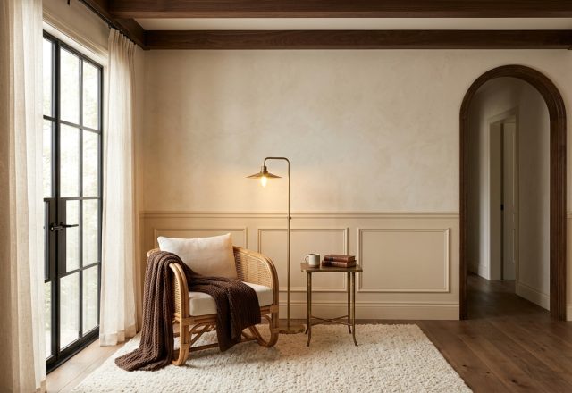 A warm neutral living room featuring textured cream walls, a rattan armchair with a brown knit throw, and an arched hallway.