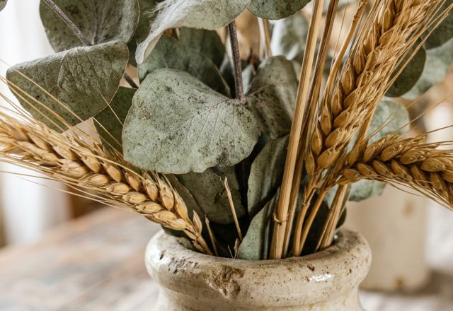 Dried eucalyptus and wheat decor arranged inside a rustic speckled ceramic vase on a wooden tabletop.