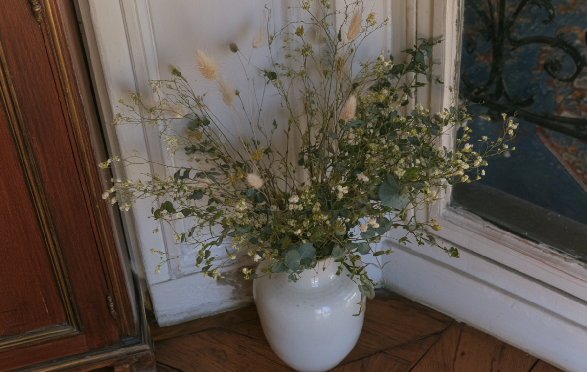 Tall dried floral arrangements in a white ceramic vase resting on a wood floor near a paneled wall.