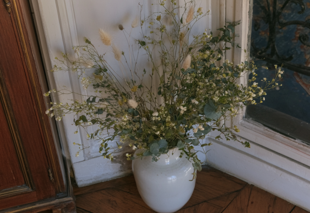 Tall dried floral arrangements in a white ceramic vase resting on a wood floor near a paneled wall.