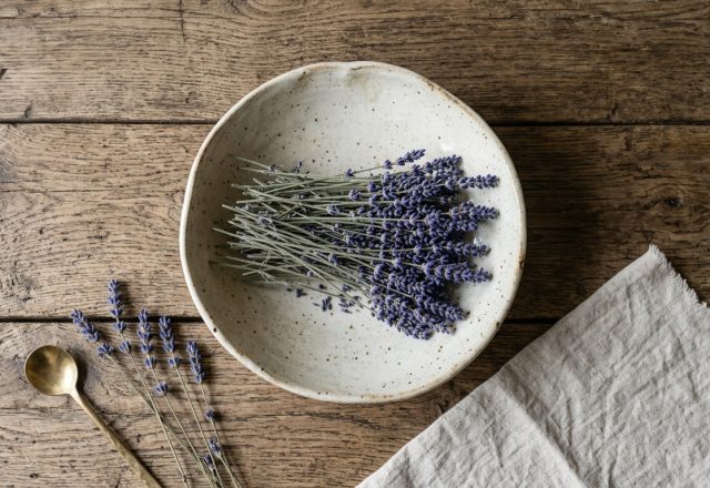 Dried lavender stems inside a speckled ceramic bowl on a rustic wooden table with linen and a brass spoon