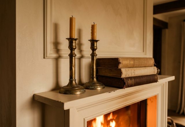 A cozy fireplace mantel styled with vintage brass candlesticks and stacked books above a warm fire.