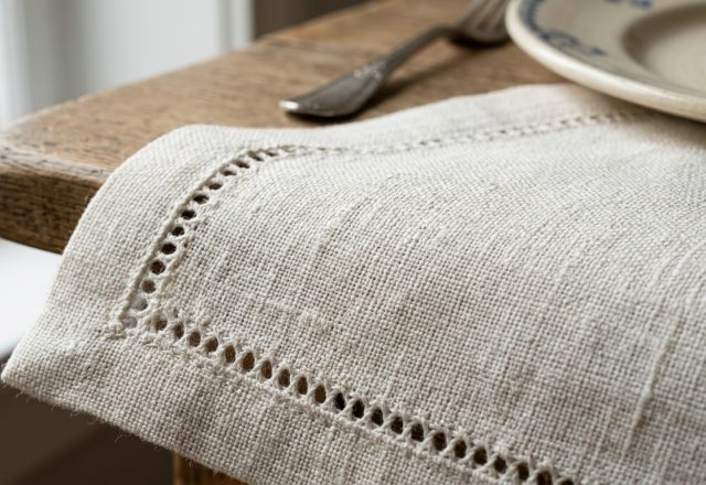 Close-up of a neutral linen napkin with hemstitch details resting on a wooden table beside a ceramic plate.