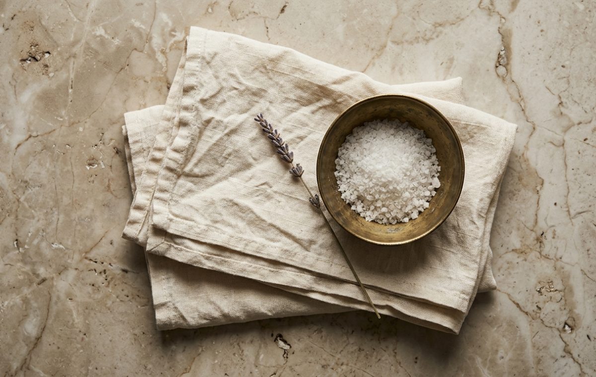 Coarse white bath salt in a brass bowl resting on a beige linen towel with dried lavender on marble.