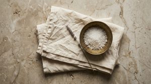Coarse white bath salt in a brass bowl resting on a beige linen towel with dried lavender on marble.