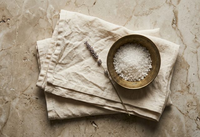 Coarse white bath salt in a brass bowl resting on a beige linen towel with dried lavender on marble.