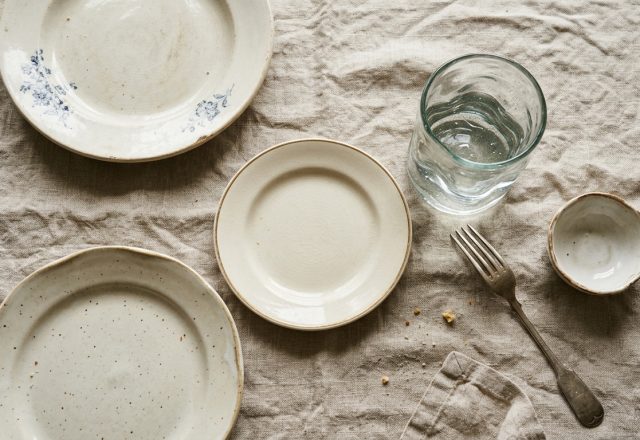 A rustic table setting featuring ceramic plates, linen tablecloth, glass of water, and a fork on neutral fabric background in a Japandi style dining space.