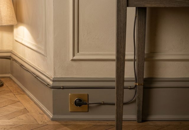 Exposed cable management using brass cable clips and fabric cord running along historic grey baseboards.