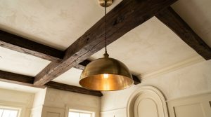Brushed brass pendant light hanging beneath dark exposed ceiling beams in a transitional interior space.