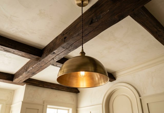 Brushed brass pendant light hanging beneath dark exposed ceiling beams in a transitional interior space.
