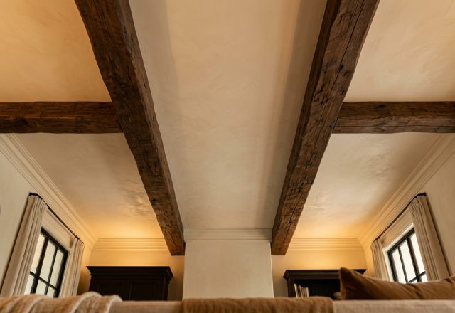 Rustic exposed ceiling beams crossing a textured plaster ceiling in a bright living space.