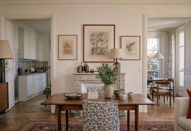 Bright French apartment dining room featuring a rustic wooden table, marble fireplace, and herringbone parquet floors.