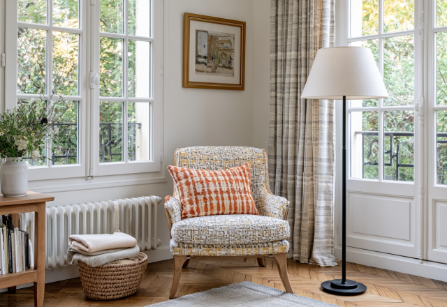Bright French Country corner seating featuring a patterned armchair, floor lamp, and wooden console by casement windows.
