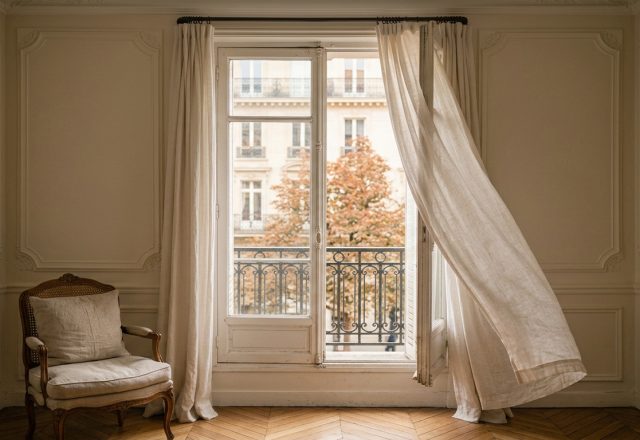 Bright French country living room featuring chevron hardwood floors, linen curtains blowing in the wind, and vintage armchair.