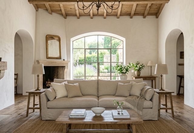 Bright French Country living room featuring exposed wood beams, a stone fireplace, arched windows, and neutral linen sofa.