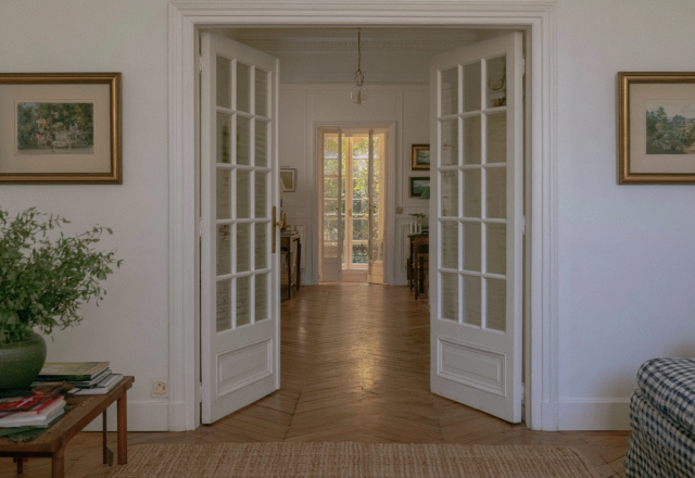 Classic white French doors open to reveal a sunlit interior passageway with herringbone wood floors.