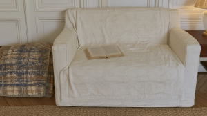 A cozy reading nook setup featuring a neutral slipcovered armchair, an open book, and a textured plaid blanket.