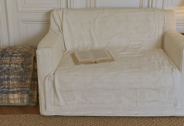 A cozy reading nook setup featuring a neutral slipcovered armchair, an open book, and a textured plaid blanket.