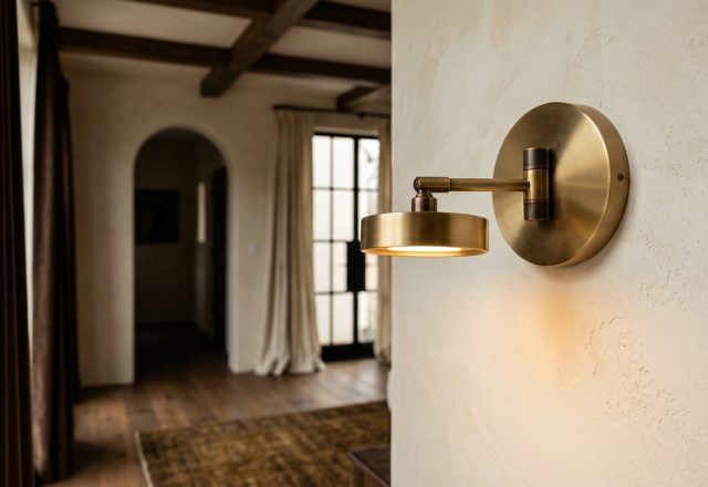 Brushed brass wall sconce casting a warm glow on a textured plaster wall in a transitional hallway.