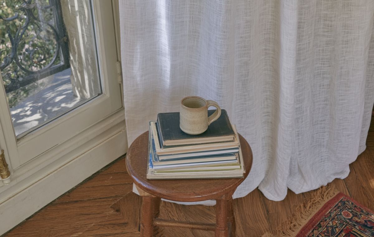 A minimalist reading nook setup featuring a wooden stool, a stack of books, and a ceramic mug near a sunlit window.