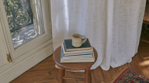 A minimalist reading nook setup featuring a wooden stool, a stack of books, and a ceramic mug near a sunlit window.
