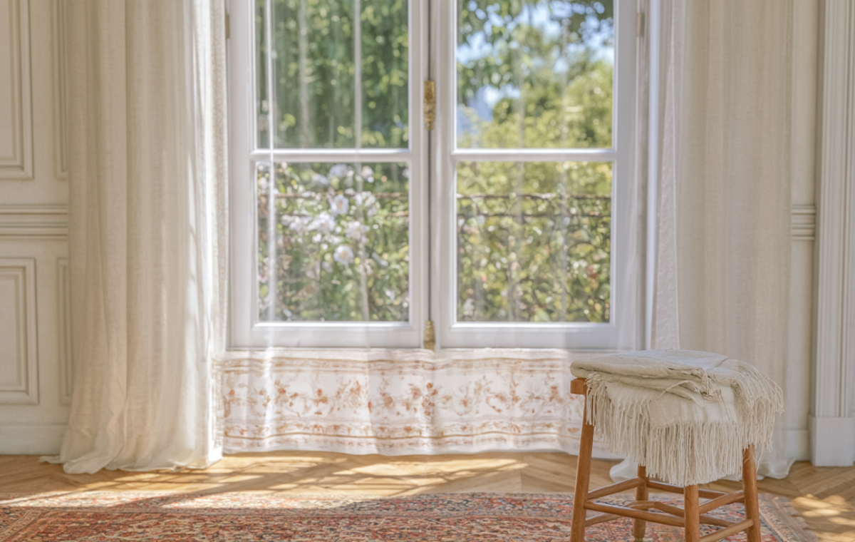 A bright natural light living room featuring sheer curtains and a wooden stool on herringbone floors.