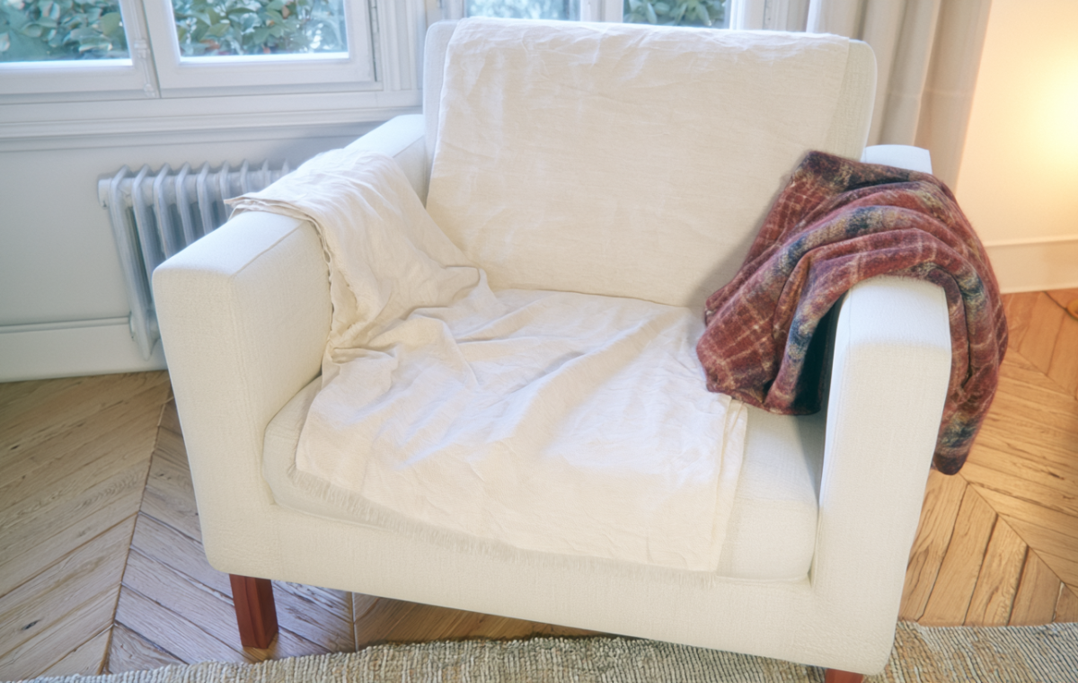 A cozy reading chair draped with a neutral linen throw and a plaid wool blanket in a bright room.
