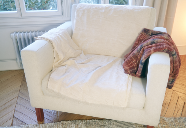A cozy reading chair draped with a neutral linen throw and a plaid wool blanket in a bright room.