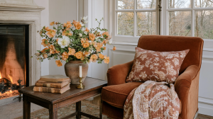 Cozy fireplace seating area featuring a rust velvet armchair, antique wooden table, and fresh autumn floral arrangement.