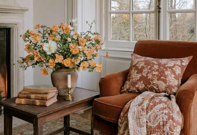 Cozy fireplace seating area featuring a rust velvet armchair, antique wooden table, and fresh autumn floral arrangement.