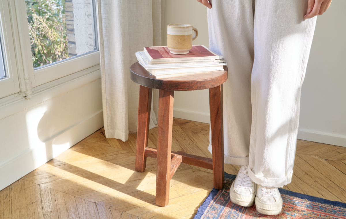 A minimalist wooden stool holding books and a coffee mug in a sunlit living room corner.
