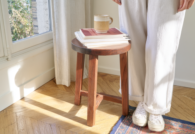 A minimalist wooden stool holding books and a coffee mug in a sunlit living room corner.