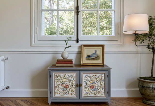 A grey painted console cabinet with floral doors sits beneath a multi-pane window with a framed bird print and ceramic vase on top.