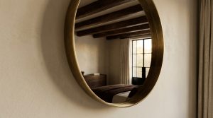 A round brass wall mirror hanging on a textured beige wall reflecting a bedroom with wooden beams