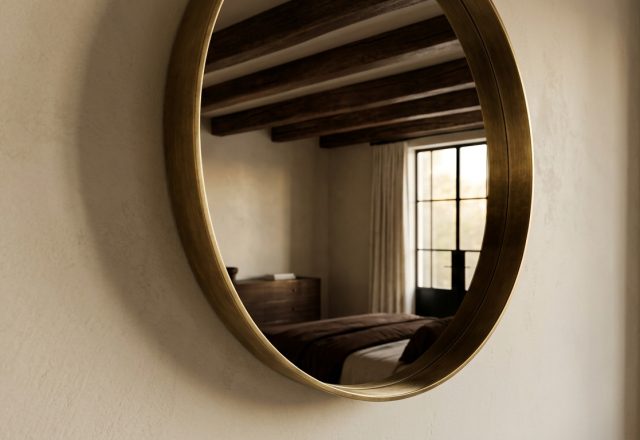 A round brass wall mirror hanging on a textured beige wall reflecting a bedroom with wooden beams