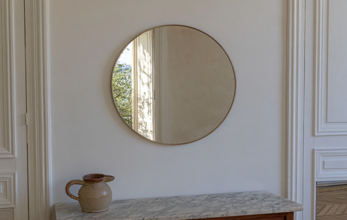 A minimalist round brass wall mirror mounted above a marble console table with an antique ceramic jug.