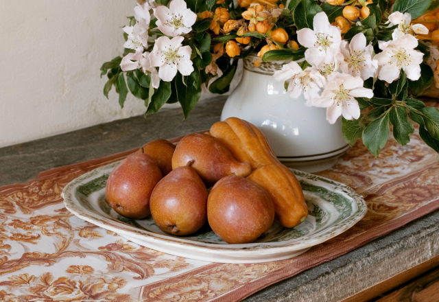 A rustic kitchen table centerpiece featuring pears and a squash on a vintage platter next to a pitcher of white blossoms.