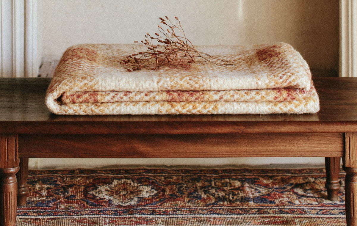 A folded vintage wool blanket and dried stems resting on a rustic wooden bench over an antique rug