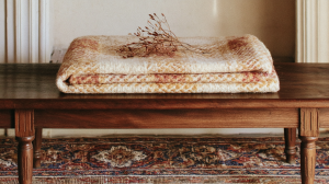 A folded vintage wool blanket and dried stems resting on a rustic wooden bench over an antique rug
