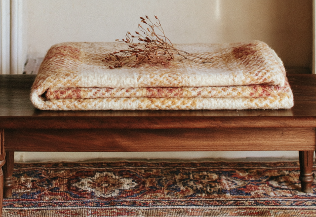 A folded vintage wool blanket and dried stems resting on a rustic wooden bench over an antique rug
