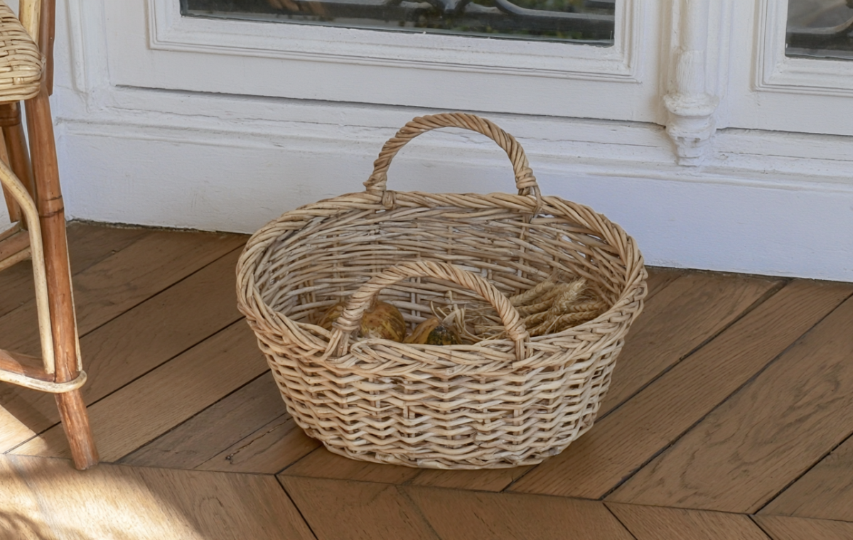 A rustic woven storage basket with double handles sitting on hardwood floors filled with dried wheat.